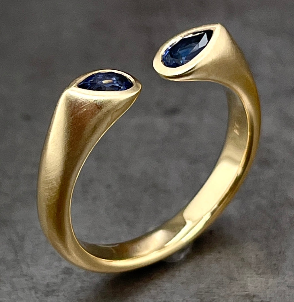 Angled aerial view of negative space pear cut sapphire ring. This ring features a yellow gold shank with a matte finish. The shank starts to come to a point in the center but leaves an opening of space before it does. On either side of the opening is a bezel set pear cut sapphire (a deep blue coloring). The sapphire is set with its round belly pointing towards the opening of the ring.