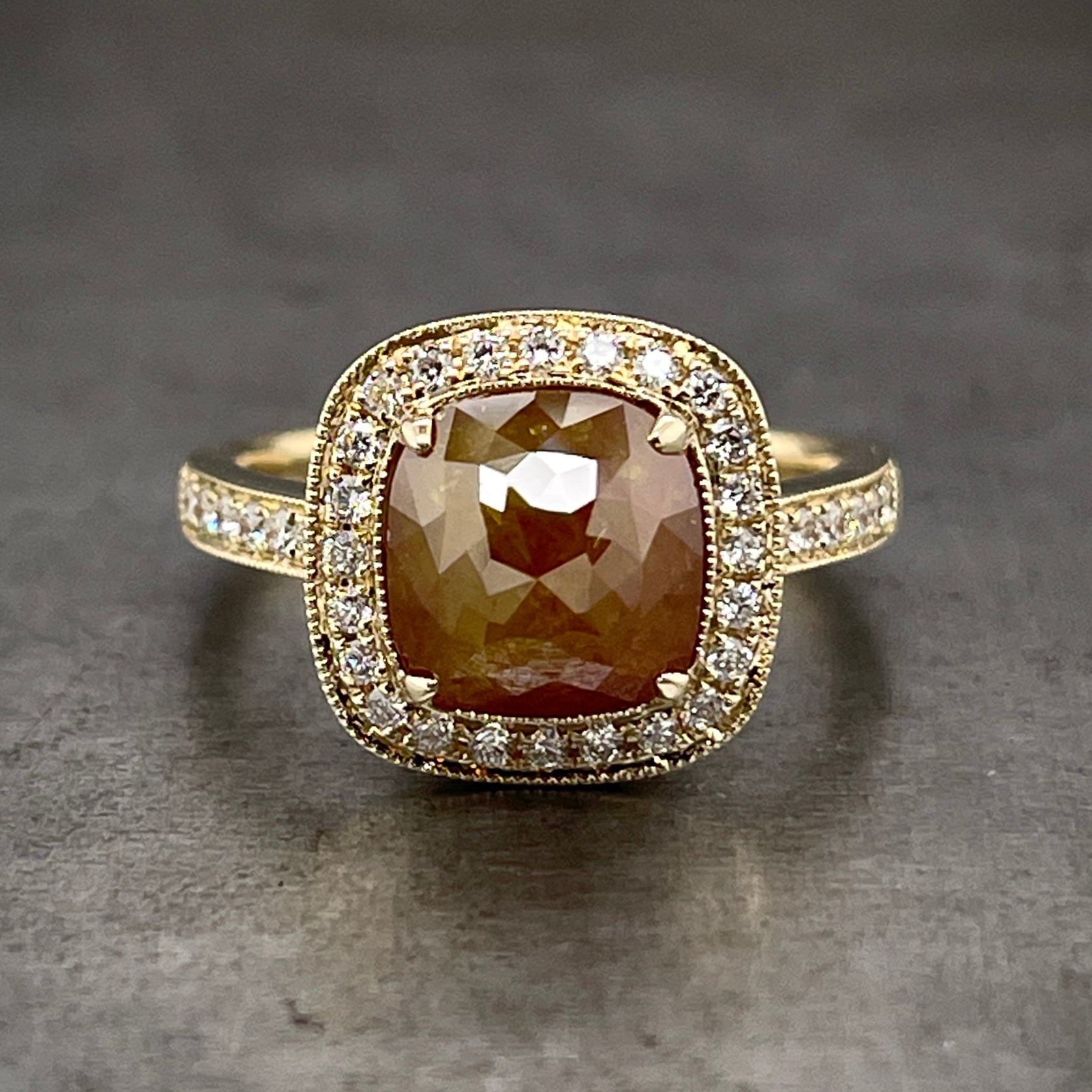 Frontal view of rose cut red-brown diamond ring. A square shaped rose cut diamond (with red, brown and yellow coloring) is four prong set in yellow gold. There is a halo of round brilliant diamonds that surrounds the rose cut diamond. There are round brilliant diamonds prong set on the shoulders of the shank. There is milgraine detailing around all the edges of the ring.