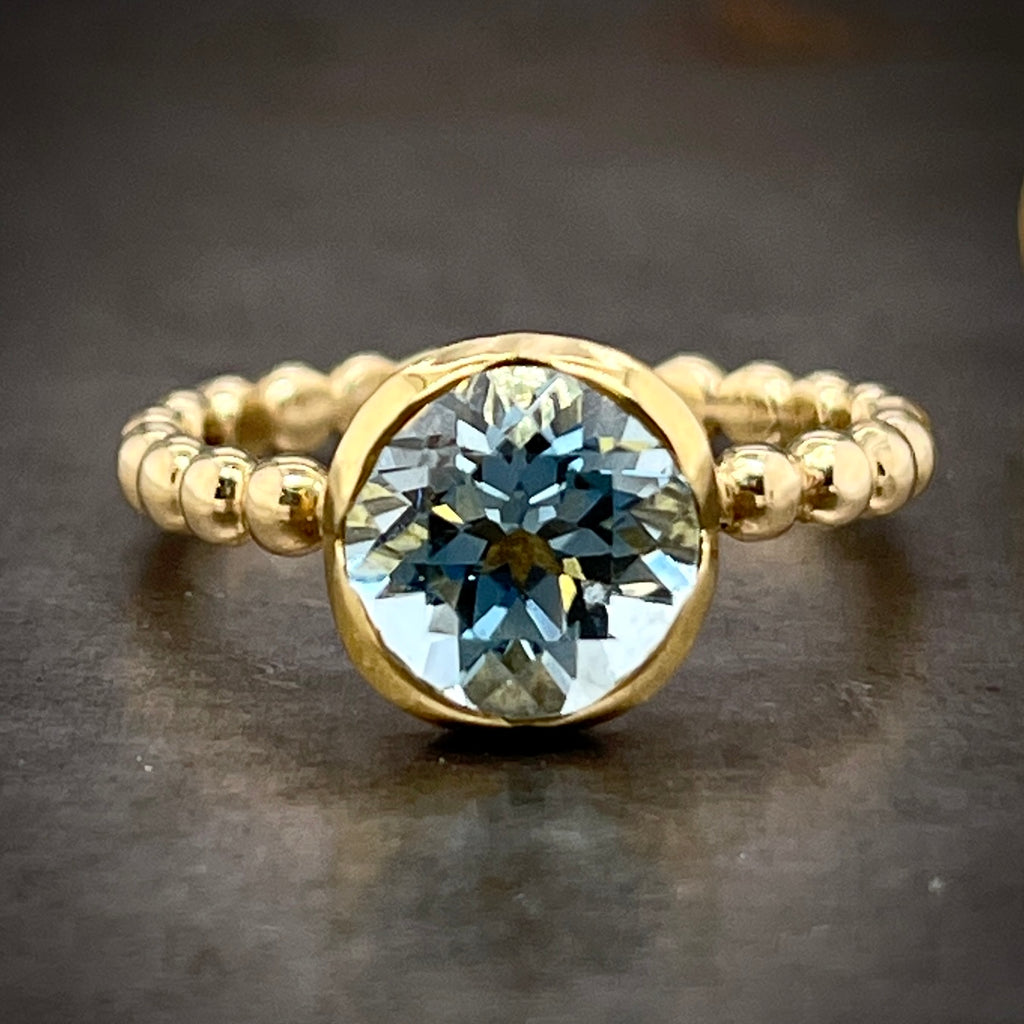 Close up view of top of ring laying down. The center of this ring features a round baby blue colored sapphire. This sapphire is bezel set in 14 karat yellow gold. The shank of this ring resembles beads made from gold.