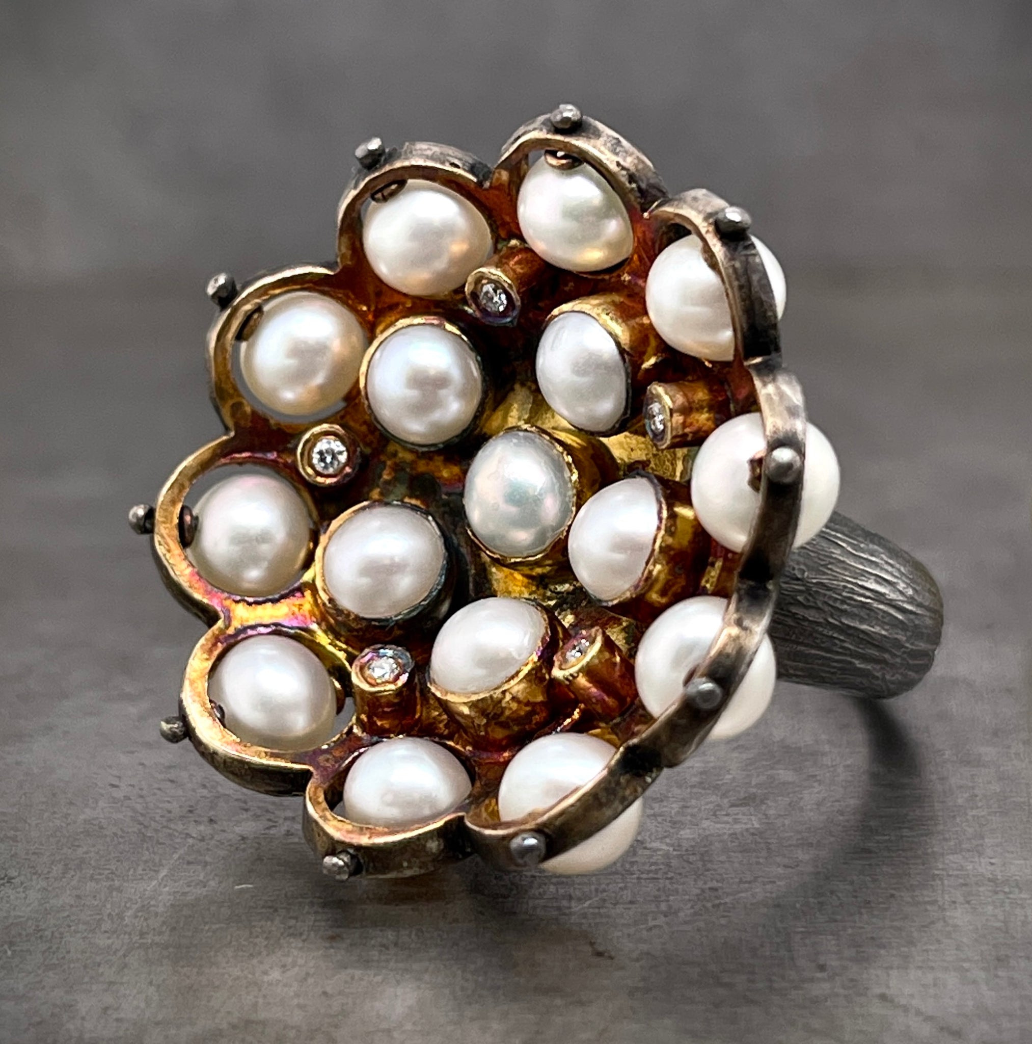 Angled side view of pearl and diamond cocktail ring. In the center there are six pearls bezel set in the formation of a flower. In between each pearl is a bezel set round brilliant diamond. The outer layer of the ring features ten pearl set on a wire so they can rotate and be moved. The inside of the ring features an oxidized yellow gold coloring, the outside is oxidized silver with a bark texture on the shank.