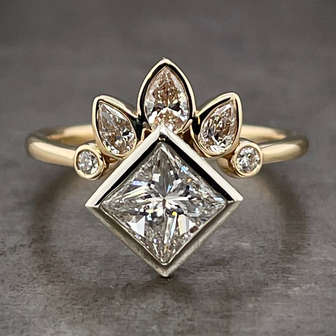 Frontal view of princess and pear cut diamond ring. A princess cut diamond is at the center, bezel set in white gold. Surrounding the top 2/5 of the ring is a tiara like diamond landscape made from pear and round diamonds. There are three pear diamonds set in the center of the tiara, with their tip pointing north (northwest and northeast for the left and right ones). Then there is one round brilliant diamond at the bottom of the tiara. They are all bezel set in yellow gold and lay on a yellow gold shank.