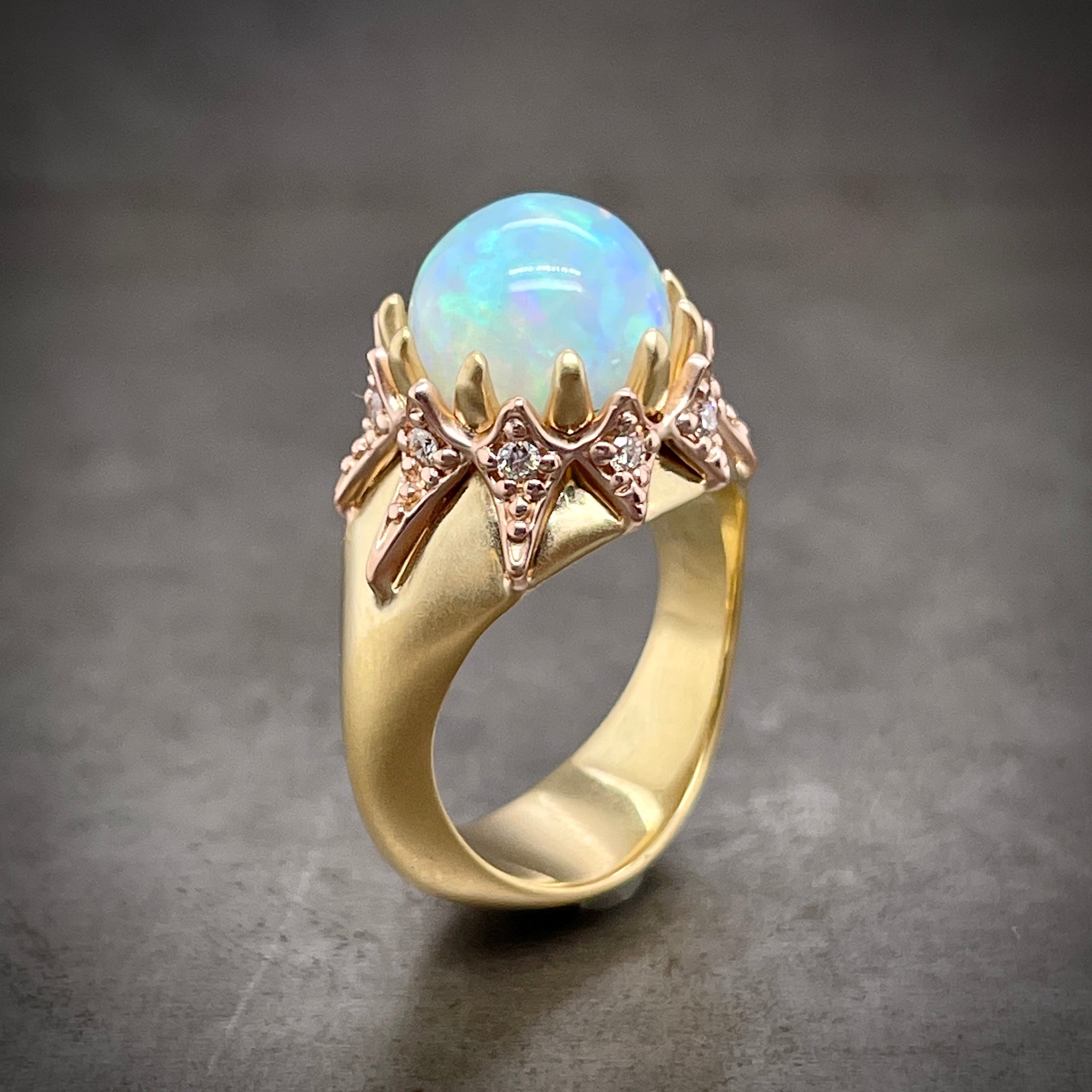 This ring features one 10mm opal claw prong set in 18 karat yellow gold. Encompassing the diameter of the prong setting is a 14 karat rose gold jacket. Within each diamond on the rose gold mounting is one round brilliant diamond. This ring features an 18 karat yellow gold shank with a matte finish.