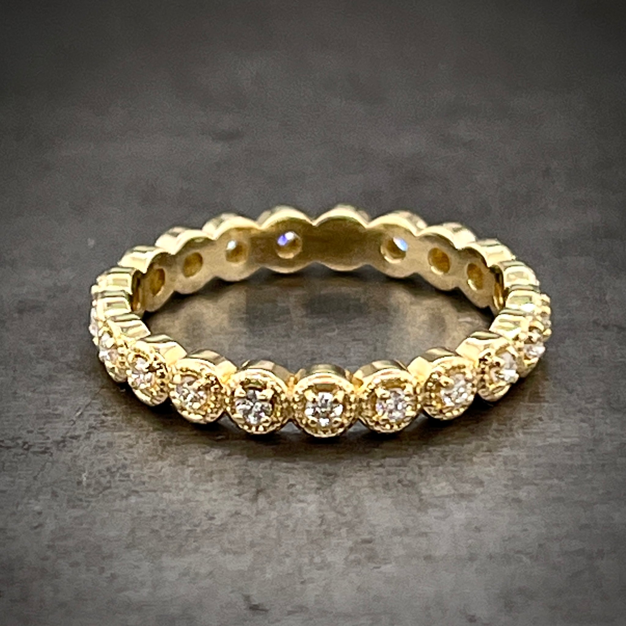 Full frontal view of diamond eternity band. This band features round brilliant diamonds that are four prong set with a curved silhouette of gold behind it that has migraine beaded detailing behind it.