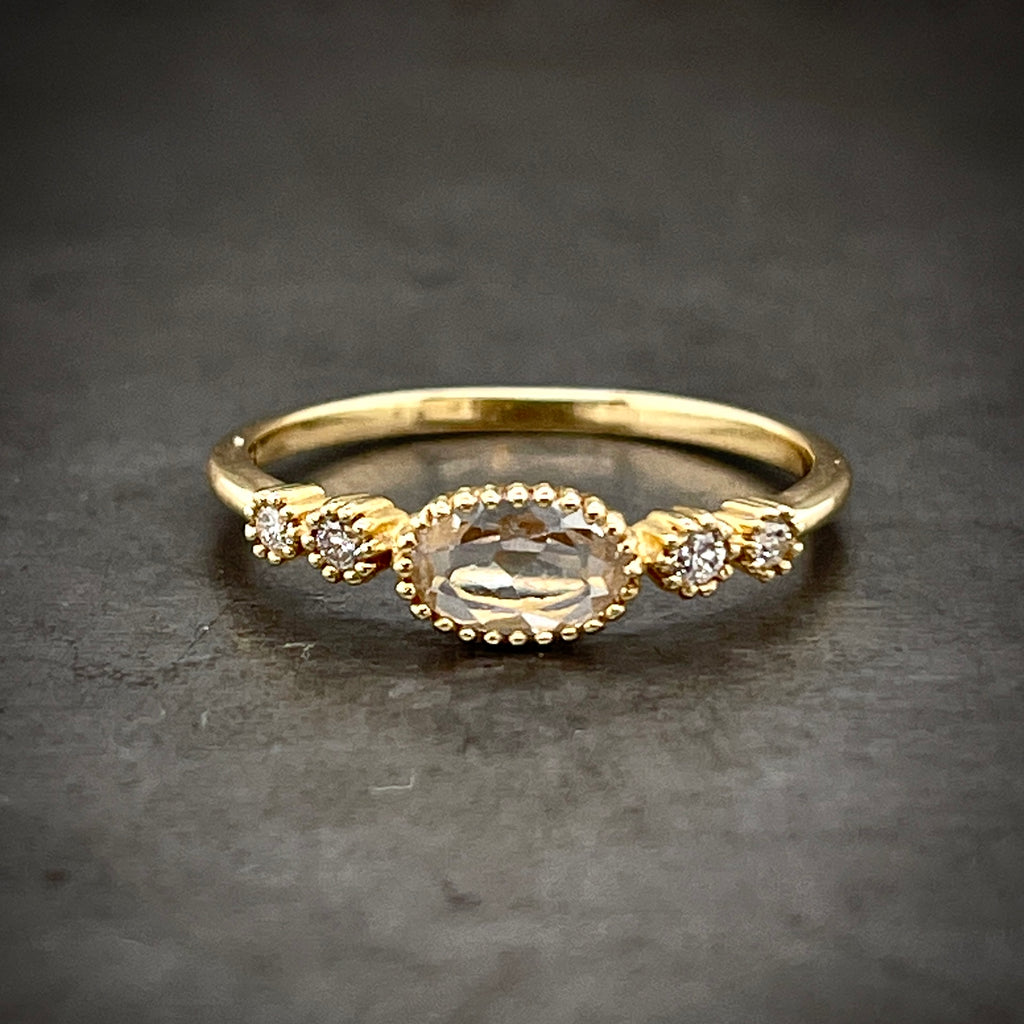 Frontal view of white topaz and diamond ring. An oval white topaz bezel set in yellow gold that has bead detailing along its boarder. Flanking either side of the topaz are two round brilliant diamonds that are bezel set in yellow gold with bead detailing.