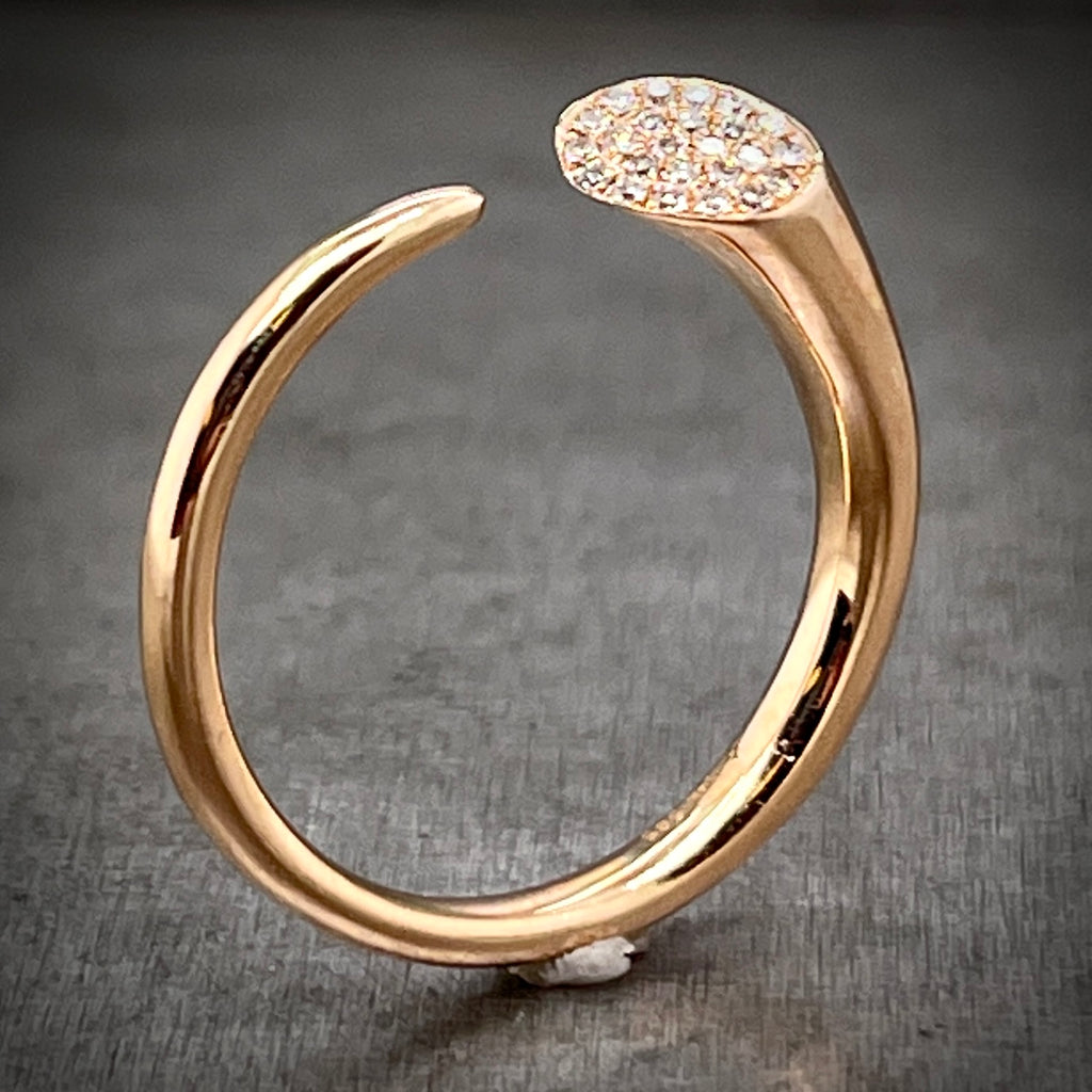 Full view of abstract rose gold and diamond ring standing up. The shank creates a perfect circle with a little space left open at the top. The left side of the shank sharpens into a point and the right becomes a thick piece of wire that is sliced at the top with round diamonds set on its face.