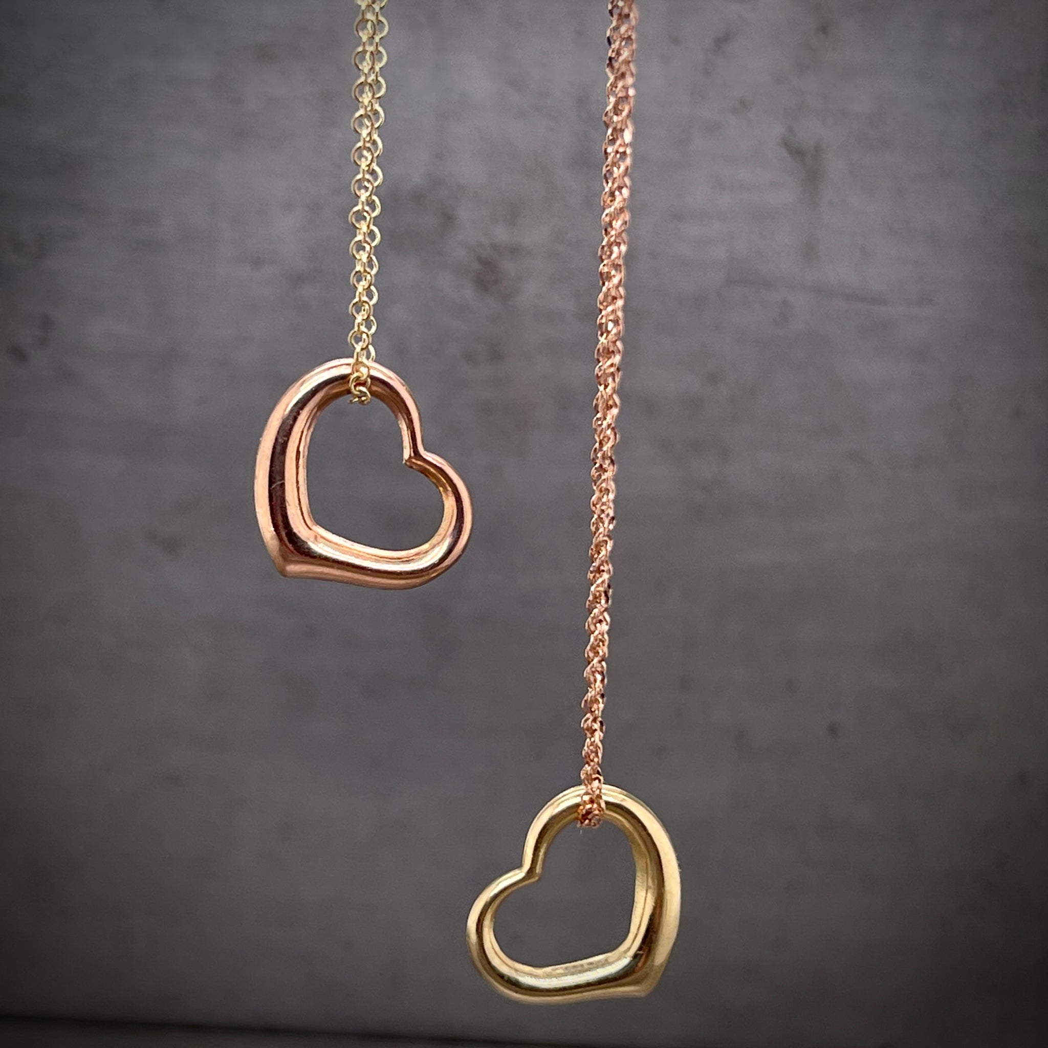 Close up image of two necklaces that each feature an asymmetrical heart pendant that slides on the chain. The necklace to the left features a yellow chain with a rose gold heart. The necklace to the right features a rose gold chain and a yellow heart.