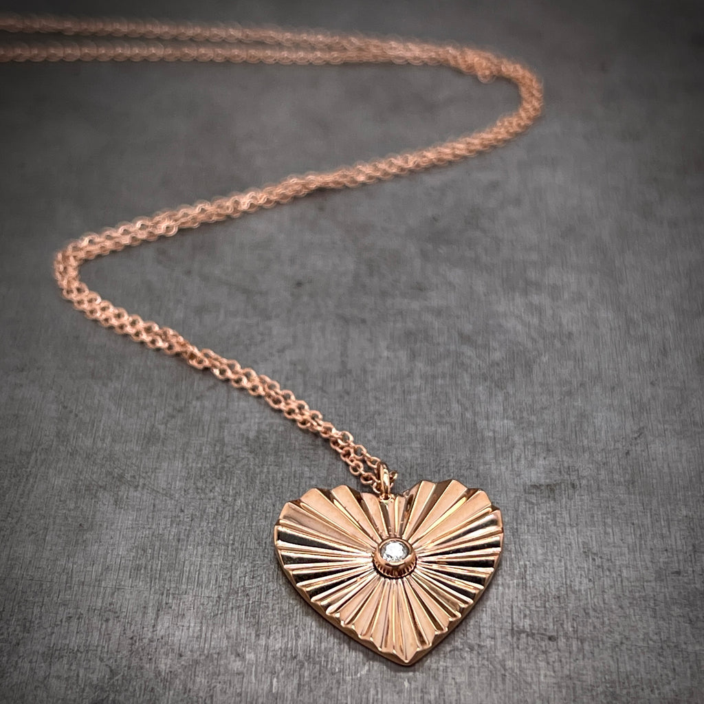 Close up view of diamond heart necklace. There is a round diamond bezel set in the center of the heart. The heart features a zig-zag motion across its surface that helps bring a focal attention to the center diamond. Rose gold pendant lays on a rose gold chain that twists around in the background.