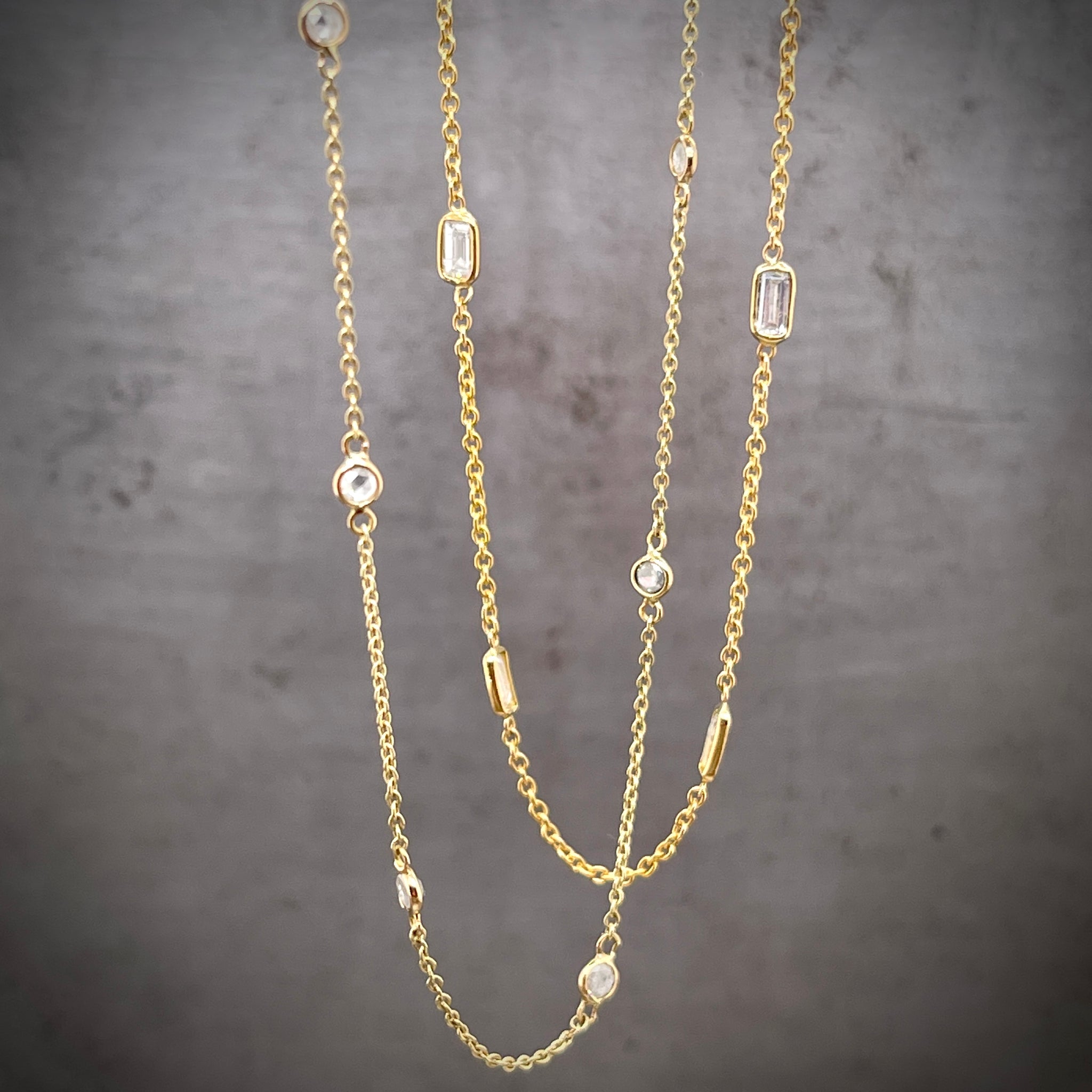 Cropped image of two necklaces hanging. The front-most necklace features round diamonds that are bezel set in gold and have an inch of chain between each diamond. The further back diamond necklace features baguette diamonds that are bezel set at every inch.