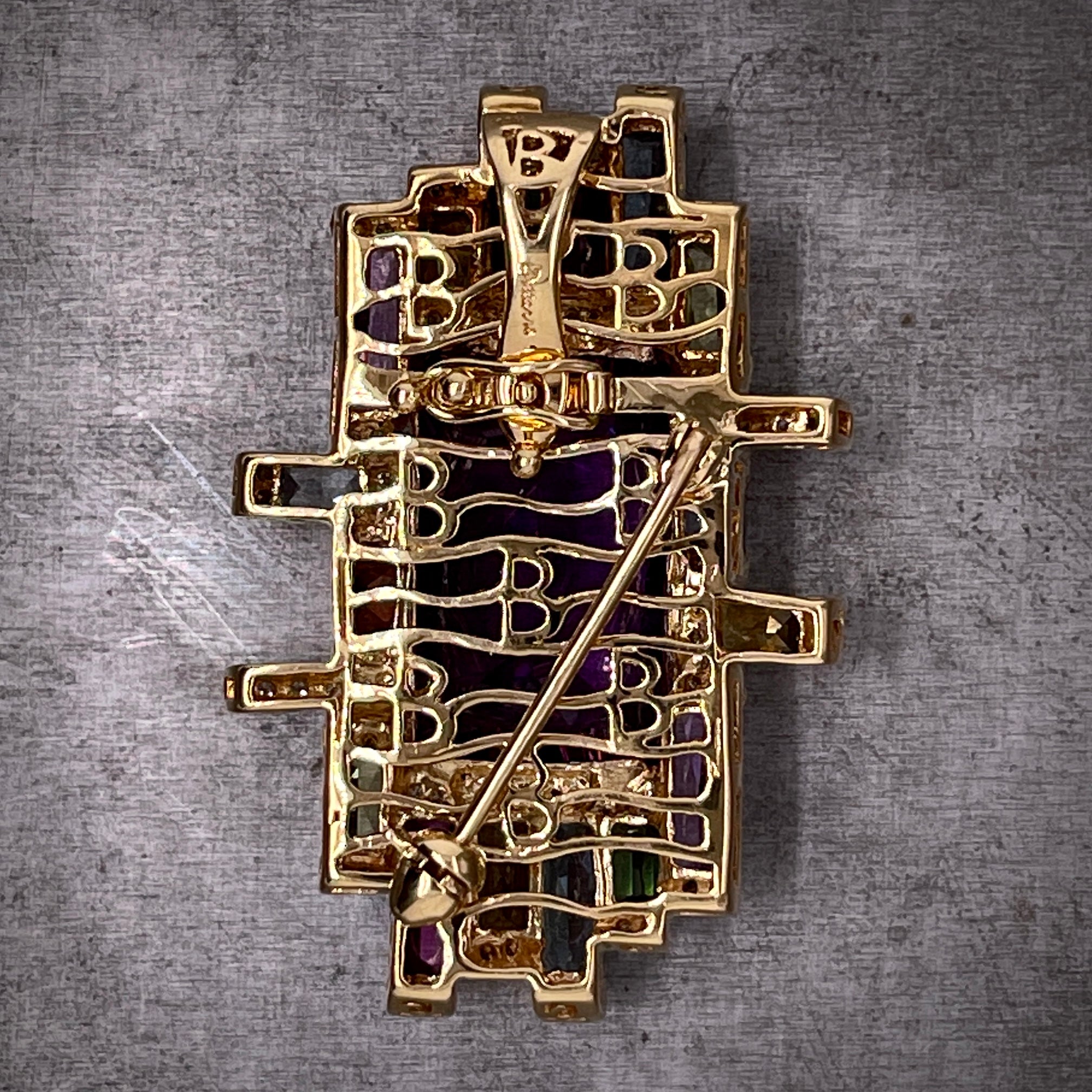 Full view of the back of the amethyst brooch. here you can see a decorative 18 karat yellow gold backing with a wavy design throughout it. There is a pin that expands from the middle right side to the bottom left. The hidden hinged bail is located at the top of the broochs' back.