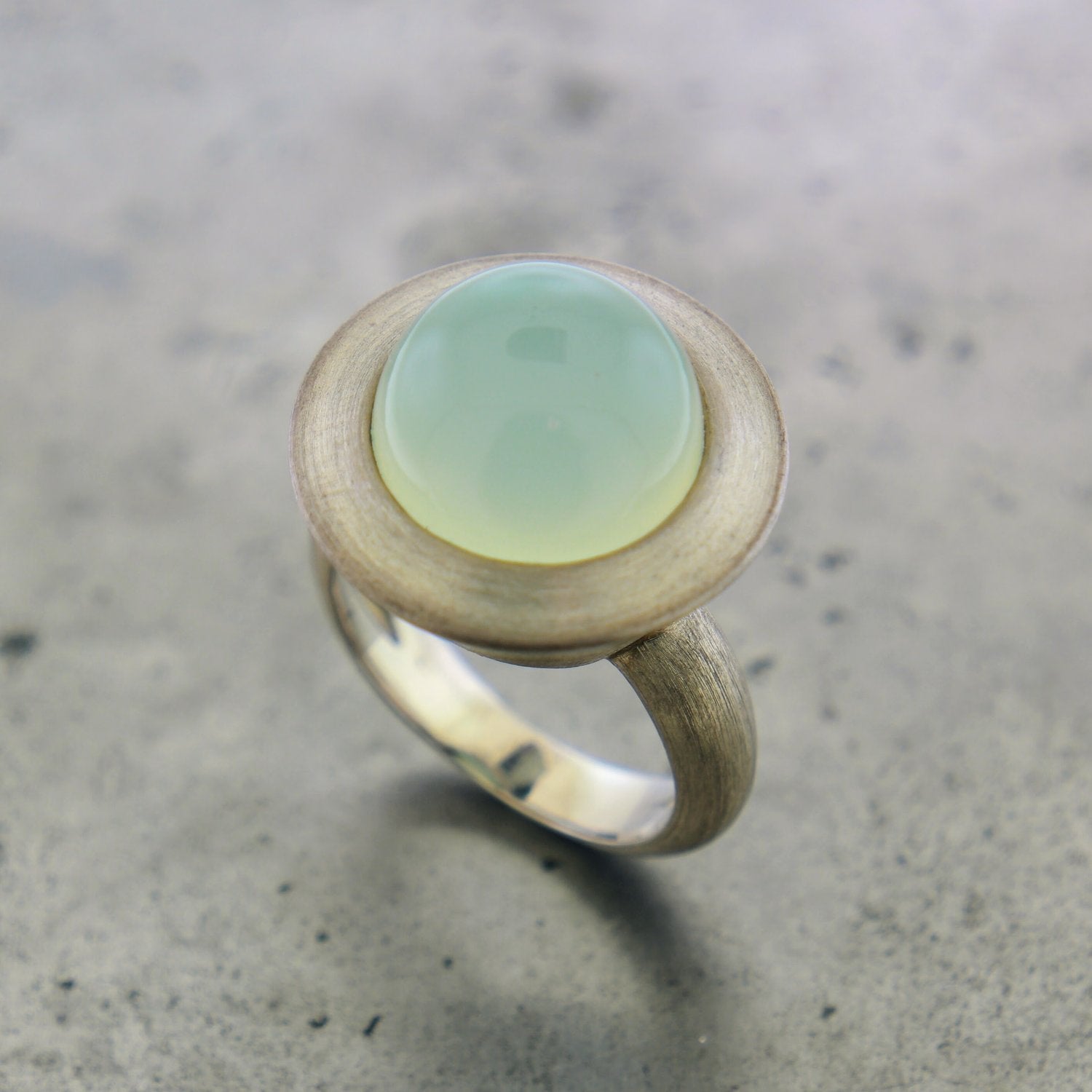 Angled aerial view of sterling silver calcedony ring. A round cabochon cut calcedony stoner (light light blue) is flush set with silver surrounding it that makes it look like an alien space ship.