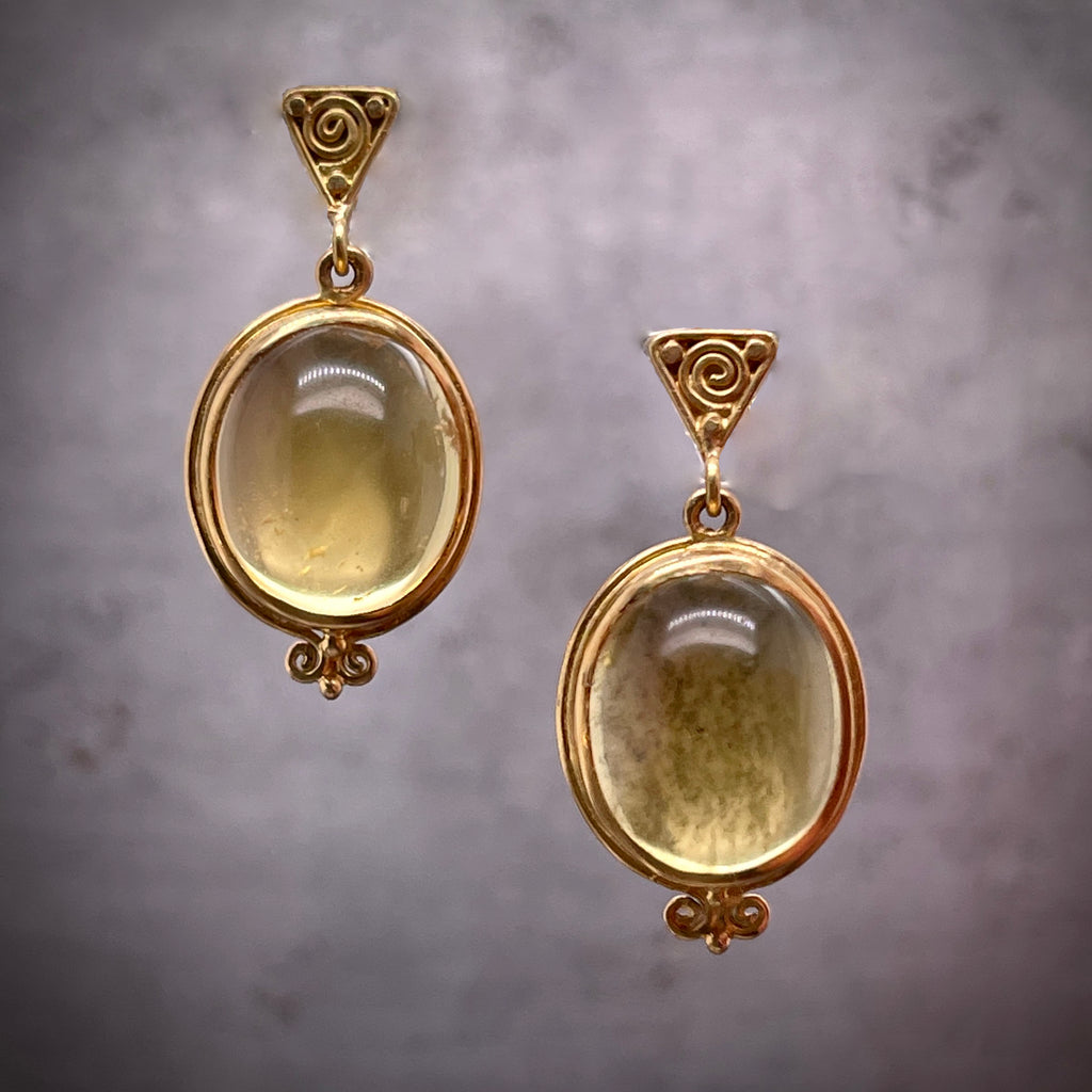 Full view of yellow tourmaline earrings hanging, the left one hangs slightly higher than the right. The stud of the earrings is a triangle that features a swirl design within it. Hanging from the triangle is an oval cut cabochon yellow tourmaline that is bezel set in yellow gold with a swirling filagree hanging off the end.