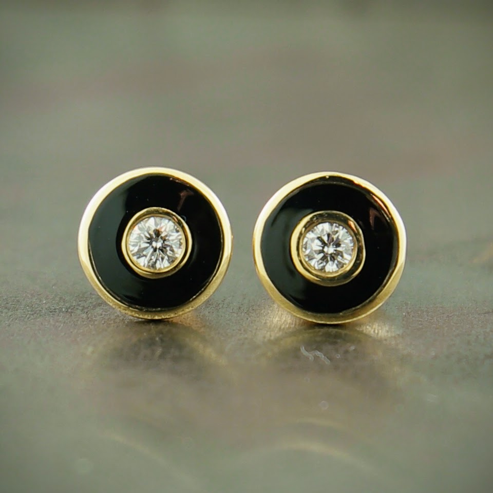 Image of 14 karat yellow gold diamond and enamel stud earrings. This image features two circle studs. In the center of these studs are a round brilliant diamond that is bezel set in 14 karat yellow gold. Surrounding the bezel setting is a layer of black enamel. Surrounding the enamel is a thin layer of 14 karat yellow gold. These earrings lay on a gray background.