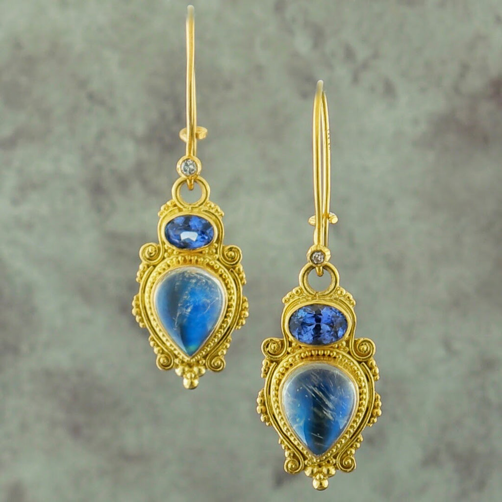 These are dangle earrings made from kidney wire. At the bottom of the wire is a small bezel set round brilliant diamond. Hanging from the diamond is a jump ring. Attached to the jump ring is a bezel set blue oval sapphire. This sapphire is oriented from West to East. Attached to the sapphire is a pear cut cabochon moonstone. This moonstone is positioned with the tip of the stone pointing South. Surrounding the sapphire and moonstone are decorative milgraine beads and swirling motifs.