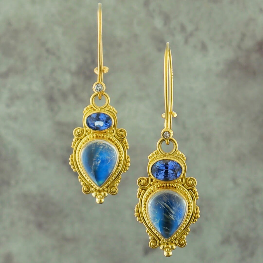 These are dangle earrings made from kidney wire. At the bottom of the wire is a small bezel set round brilliant diamond. Hanging from the diamond is a jump ring. Attached to the jump ring is a bezel set blue oval sapphire. This sapphire is oriented from West to East. Attached to the sapphire is a pear cut cabochon moonstone. This moonstone is positioned with the tip of the stone pointing South. Surrounding the sapphire and moonstone are decorative milgraine beads and swirling motifs.