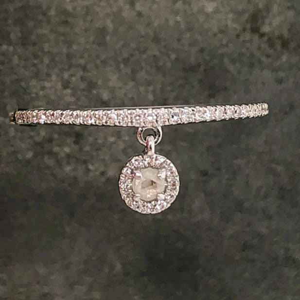 Aerial view of ring. Ring is made from 14 karat white gold and the shoulders of the shank feature one row of round brilliant diamonds prong set. In the middle of the shank, there is a jump ring layered to it with a pendant hanging from the jump ring. The pendant is of a rose-cut diamond that is four prong set in 14 karat white gold. There is a halo of round brilliant diamonds that surrounds the rose cut diamond. This pendant hangs from the ring and would move when worn. This ring lays on a gray background.