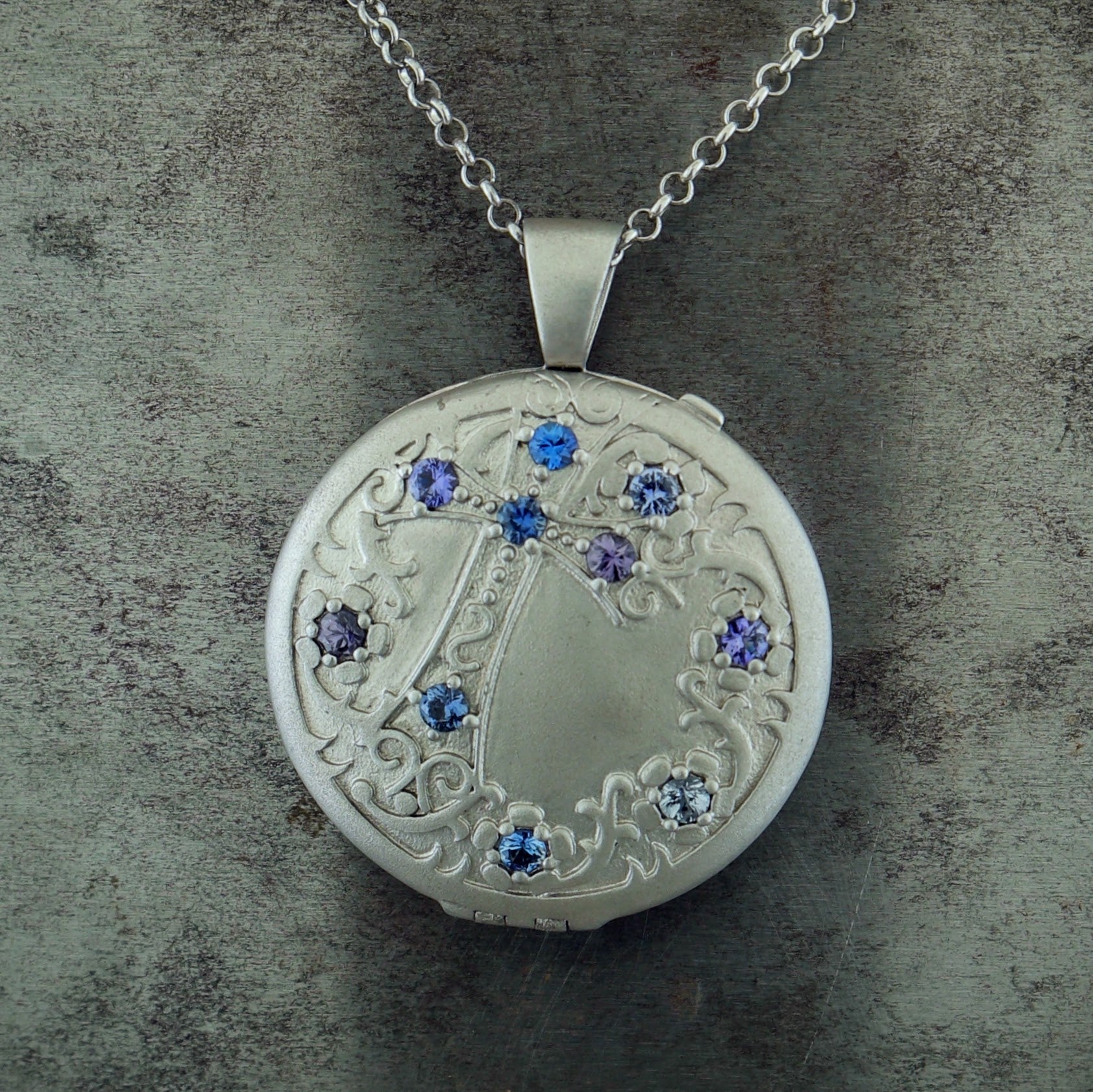 Close up view of pendant on sterling silver sapphire locket. A round locket with an engraving of a cross going diagonally across the locket with Celtic motifs surrounding the perimeter of the locket. There are ten round blue-violet sapphires set into the surface. Five of them reside within the cross; one at each end and one where all the lines intersect. The remaining five create a circle within the Celtic motifs. Pendant features a bail.