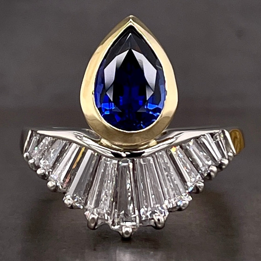 Full frontal view of pear sapphire and tapered baguette diamond ring. This ring features one blue pear sapphire positioned with its tip facing North. This sapphire is bezel set in yellow gold. Attached to the belly of the pear sapphire is a crown of tapered baguettes that are Chanel set on their northern girdles and prong set on their southern girdles.